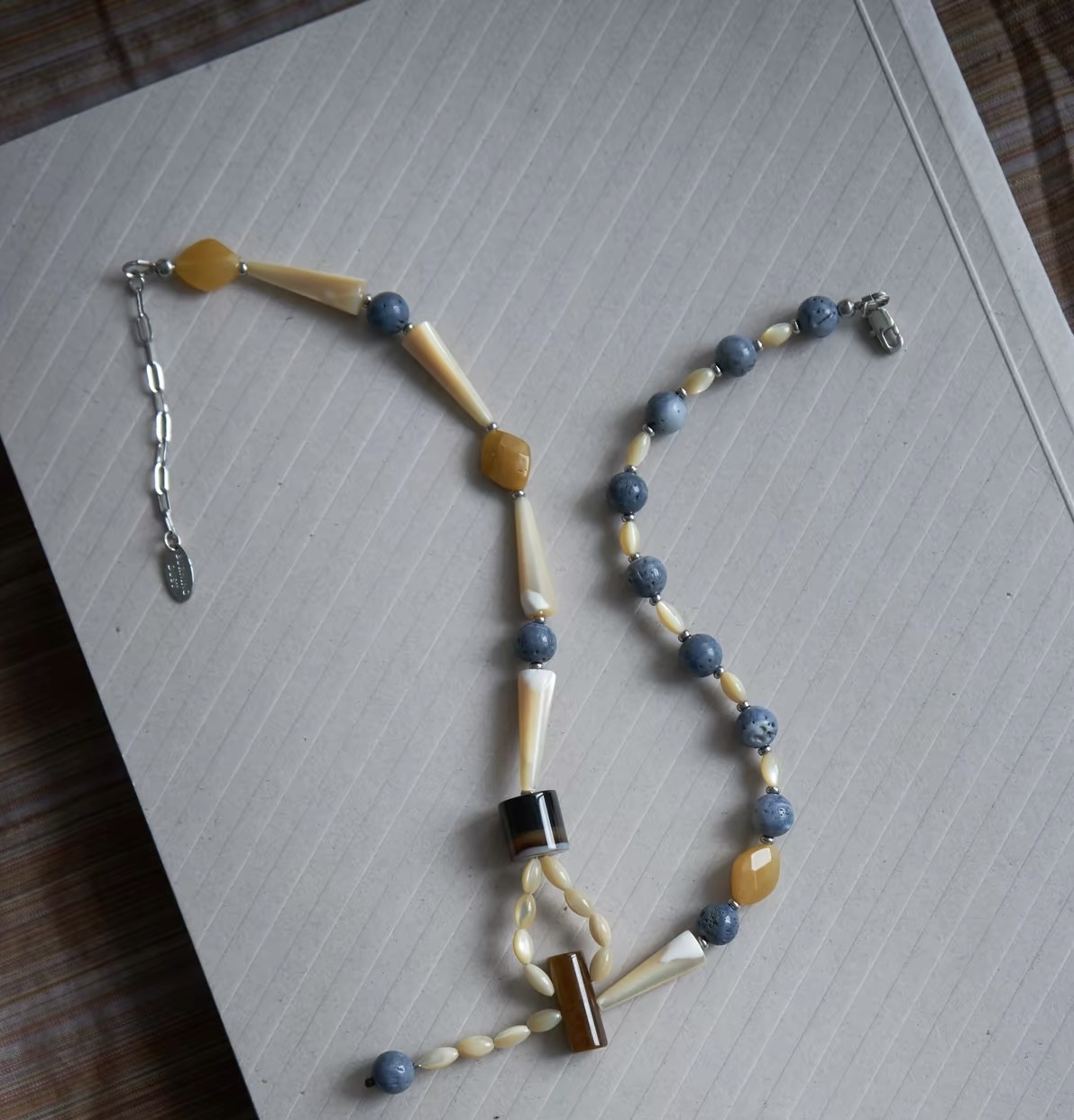 Full view of an asymmetrical natural stone and shell necklace with blue beads, yellow accents and an extension chain.