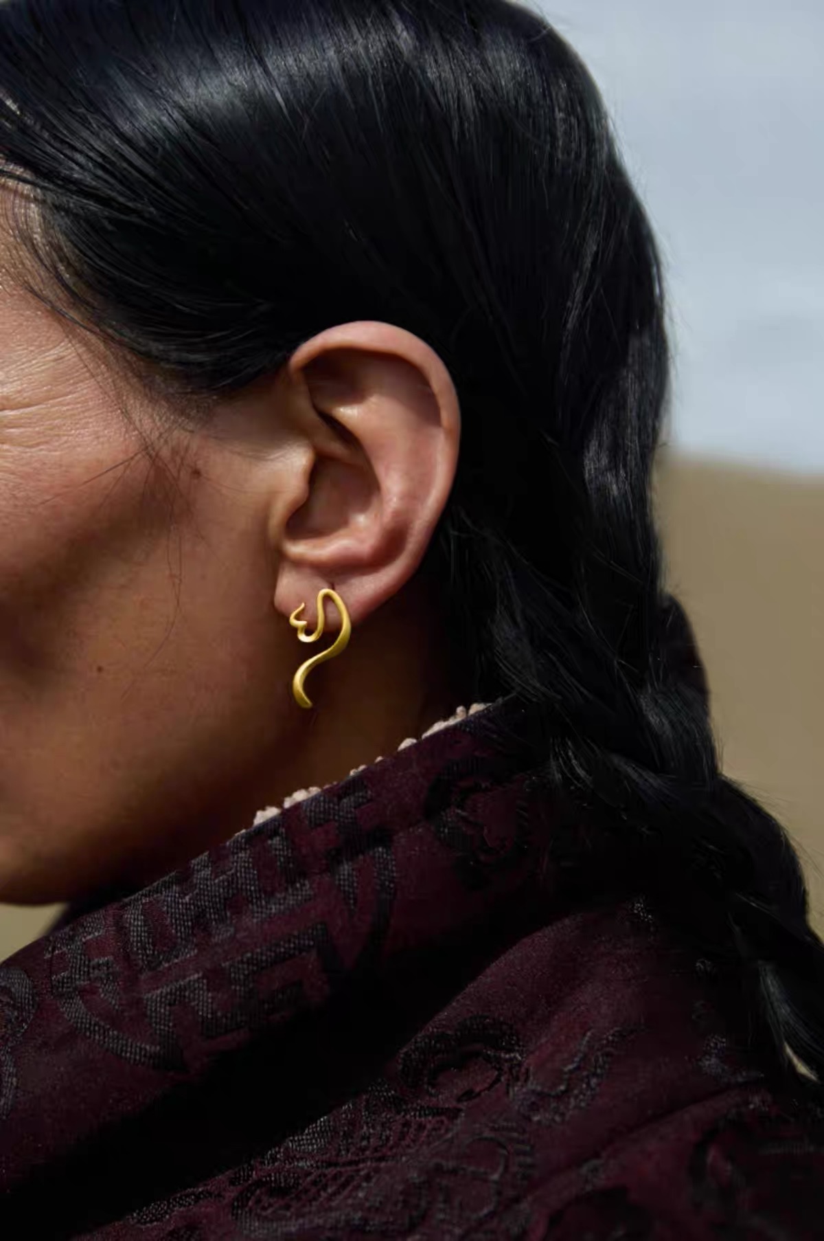 Close-up of a woman wearing a silver-tone calligraphy-inspired earring outdoors with her hair tucked behind her ear.